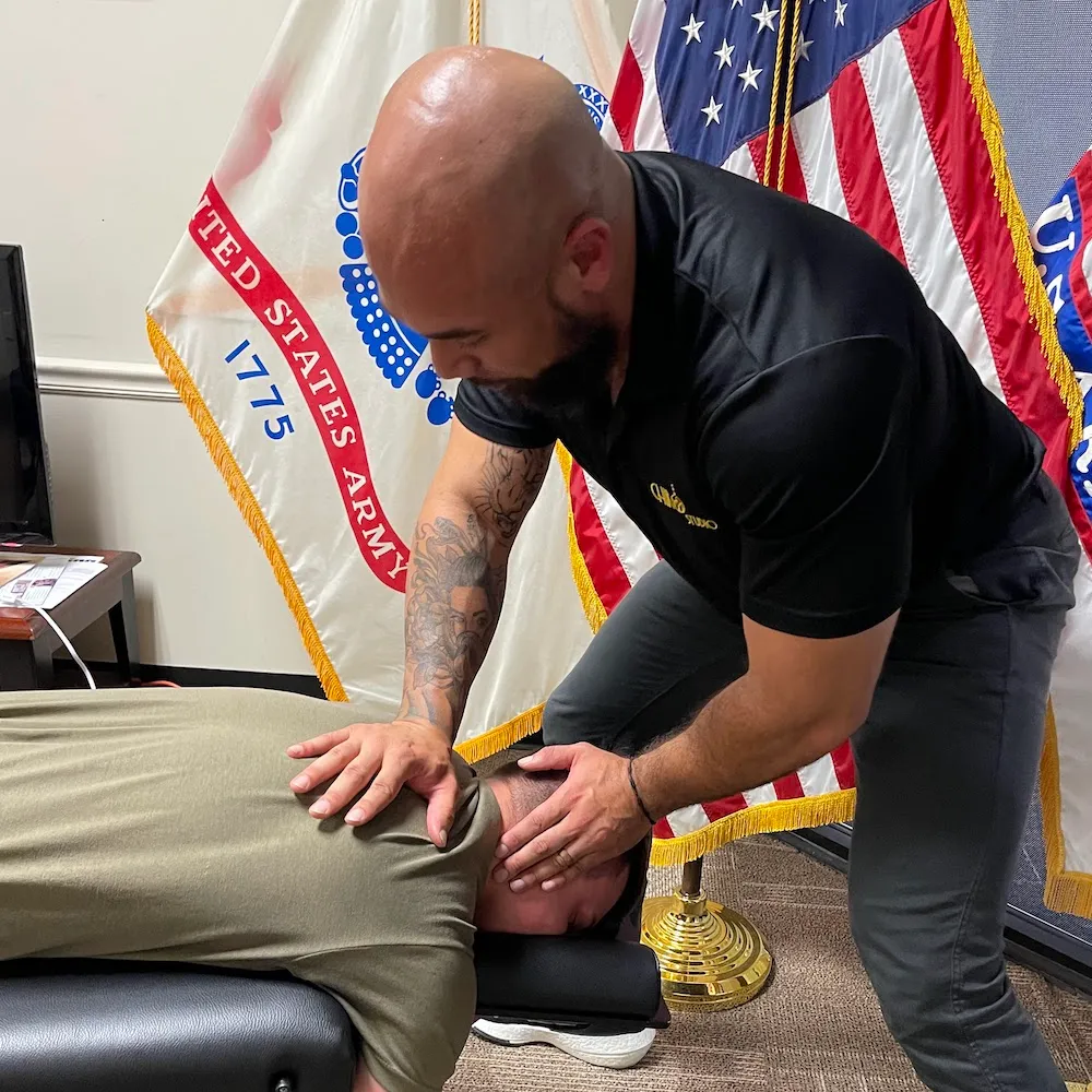 Dr. Junior adjusting the neck of a soldier lying face down, with U.S. Army and American flags behind.