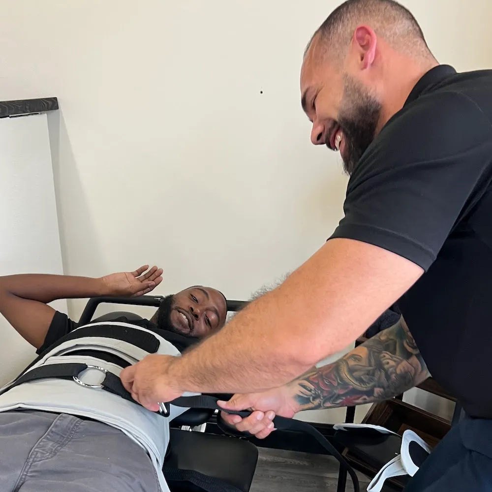 Dr. Junior fastening a decompression harness around a smiling male patient on the table.