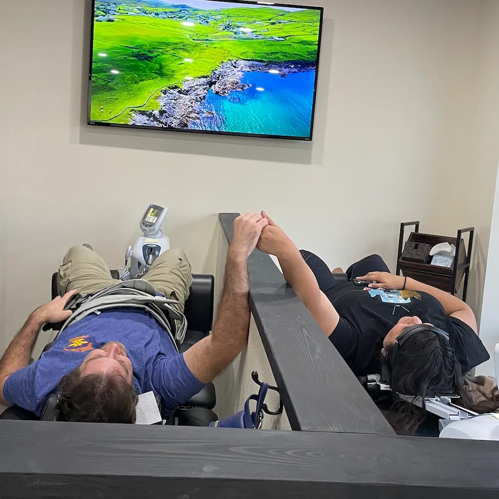 Two patients on side-by-side spinal decompression tables holding hands across the divider.