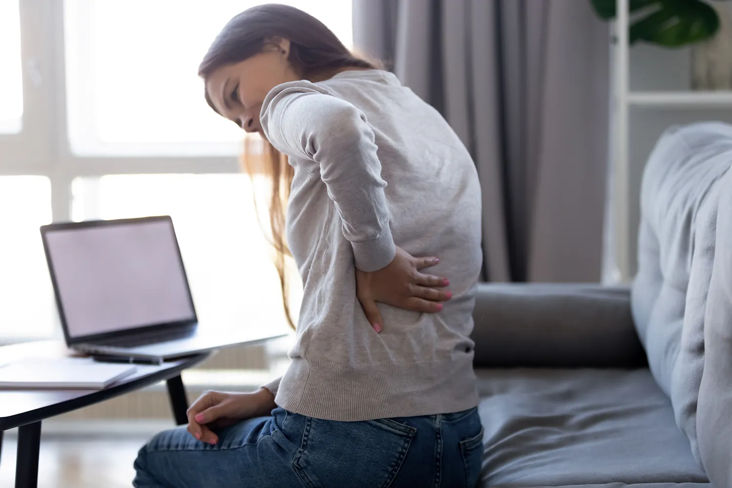 Young woman on a couch holding her lower back in pain next to a laptop.