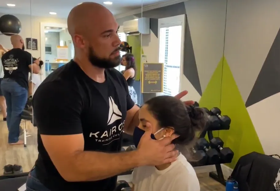 Dr. Junior adjusting the neck of a masked woman in a gym with dumbbells in the background.