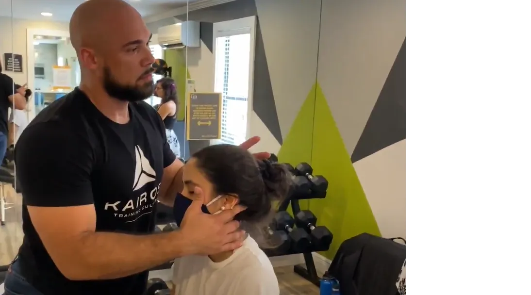 Dr. Junior adjusting the neck of a seated woman wearing a face mask in a gym.