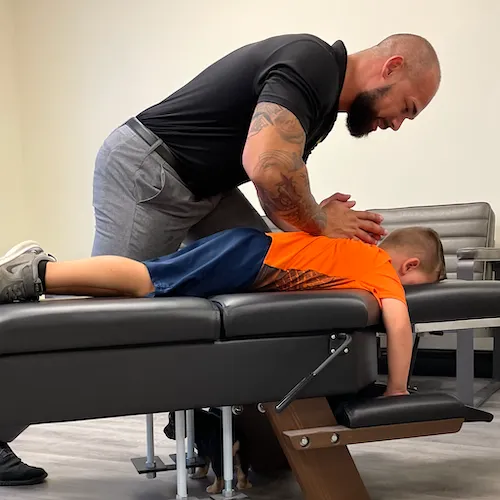 Dr. Junior smiling while adjusting a young boy lying face down on a chiropractic table.