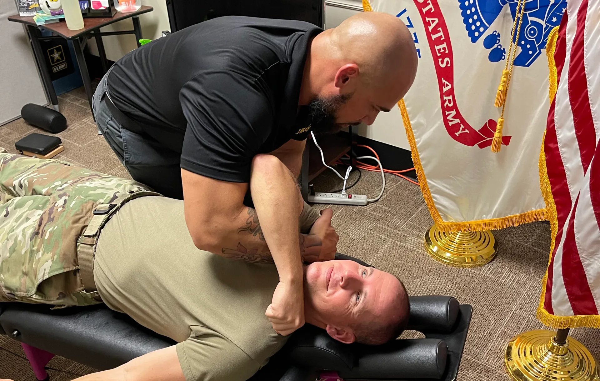 Dr. Junior performing an upper back adjustment on a soldier lying face up beside a U.S. Army flag.