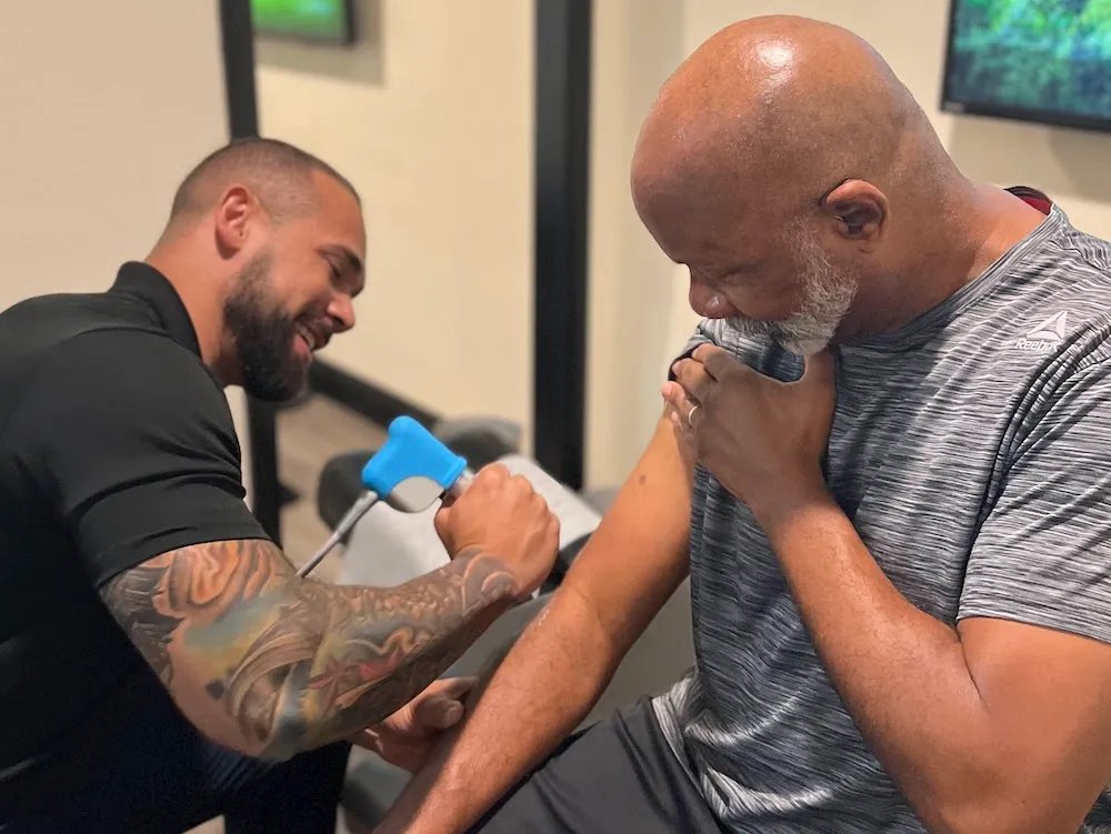 Dr. Junior smiling while treating an older man's upper arm with a shockwave therapy device.