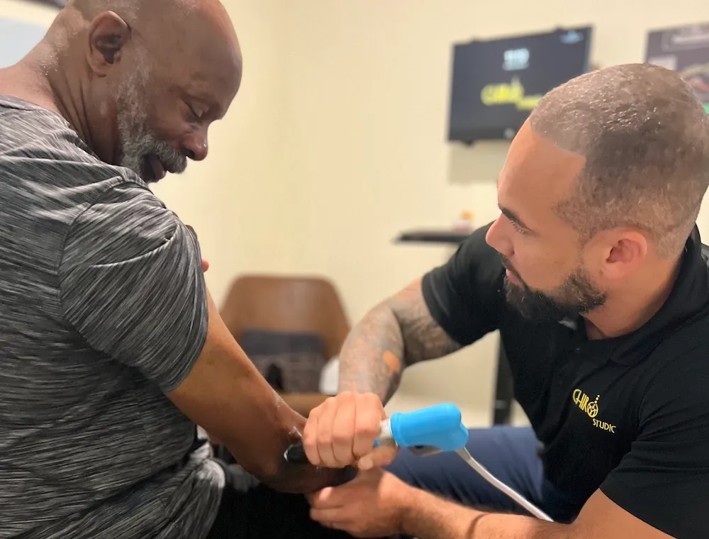 Dr. Junior focusing as he applies a shockwave therapy device to an older man's elbow.