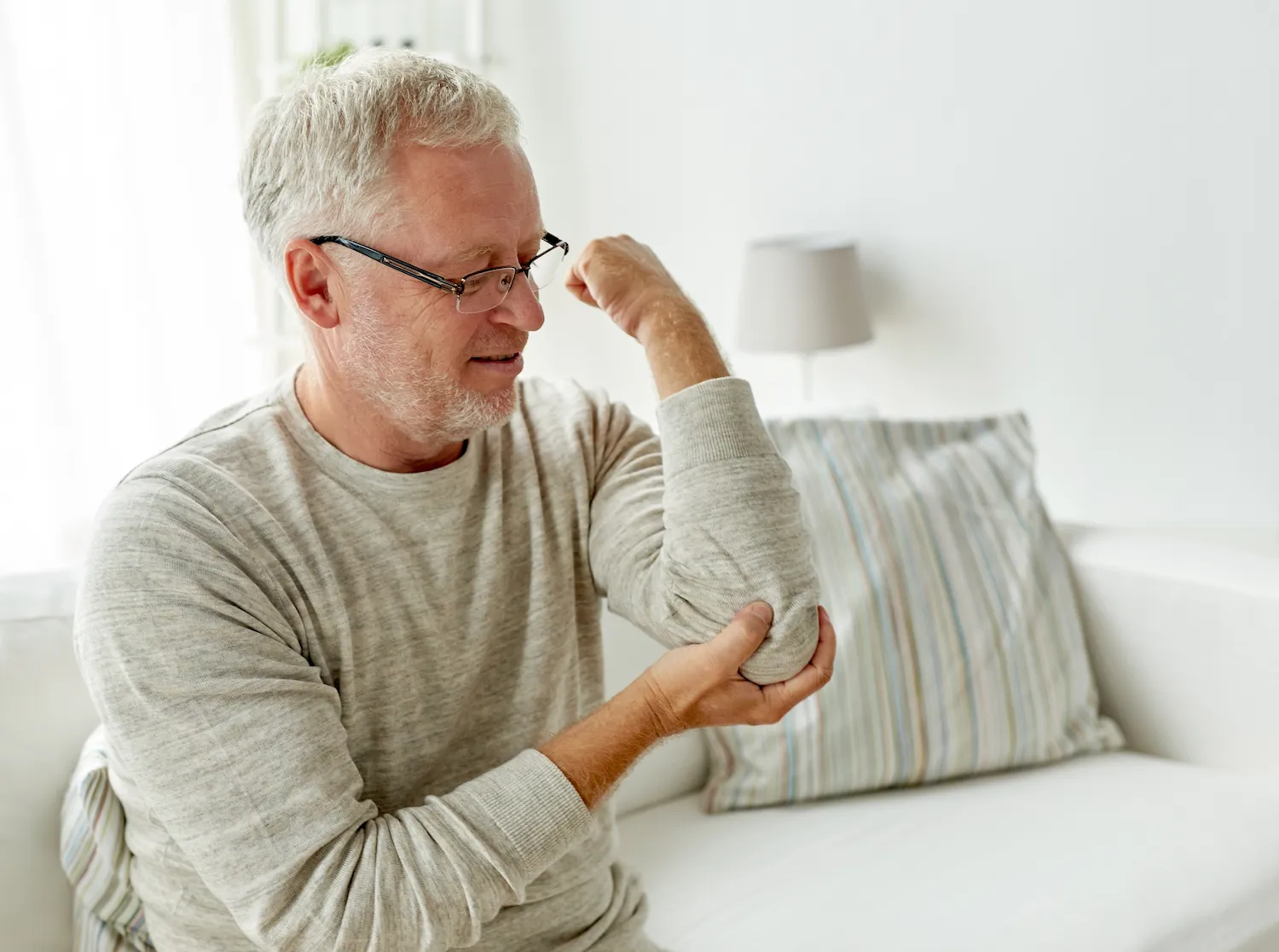 Older man in glasses sitting on a couch holding his sore elbow.