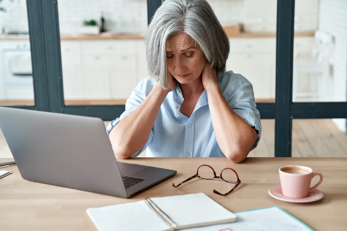 Older woman holding the back of her neck in pain while sitting at a laptop.