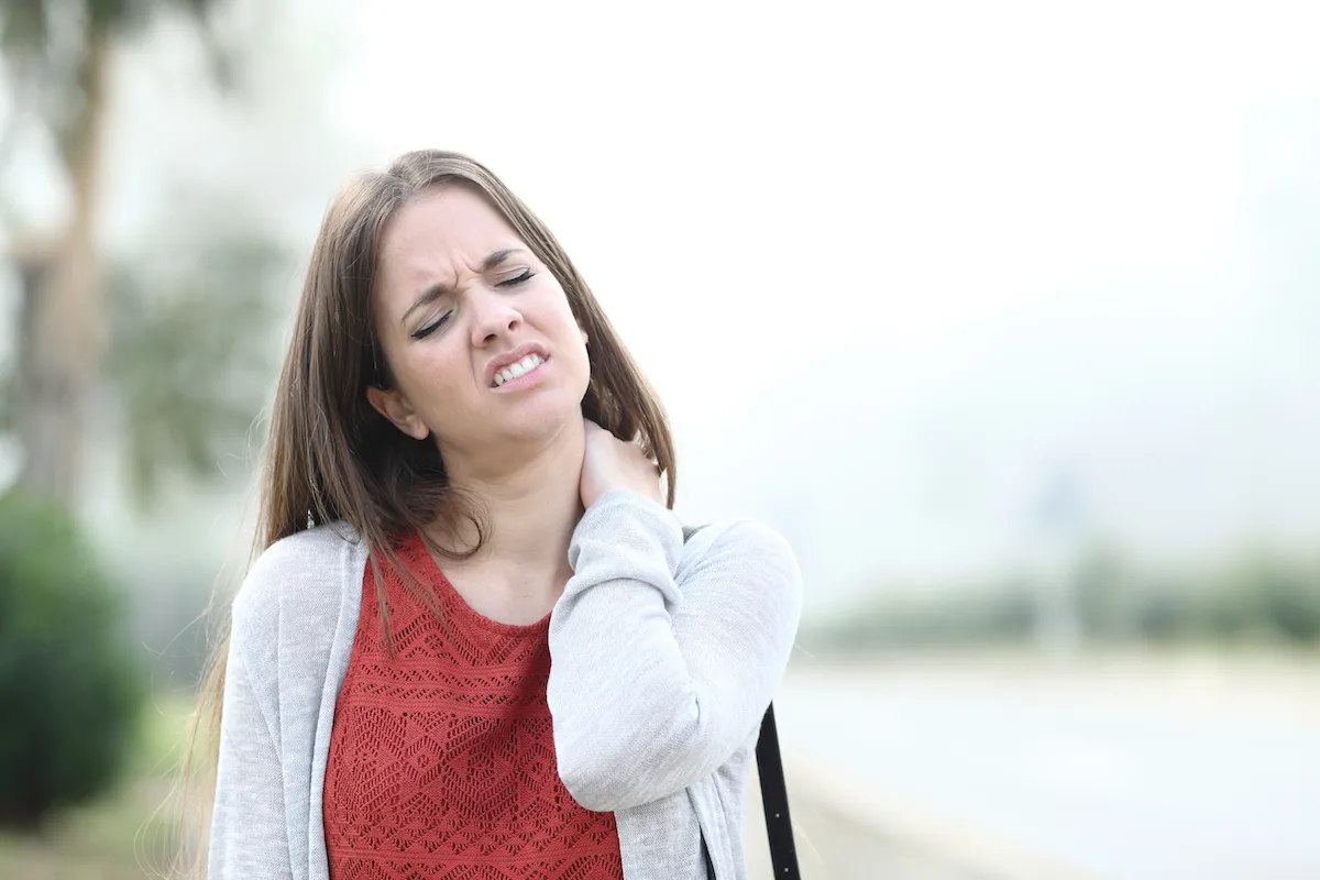Young woman wincing and holding the back of her neck outdoors.