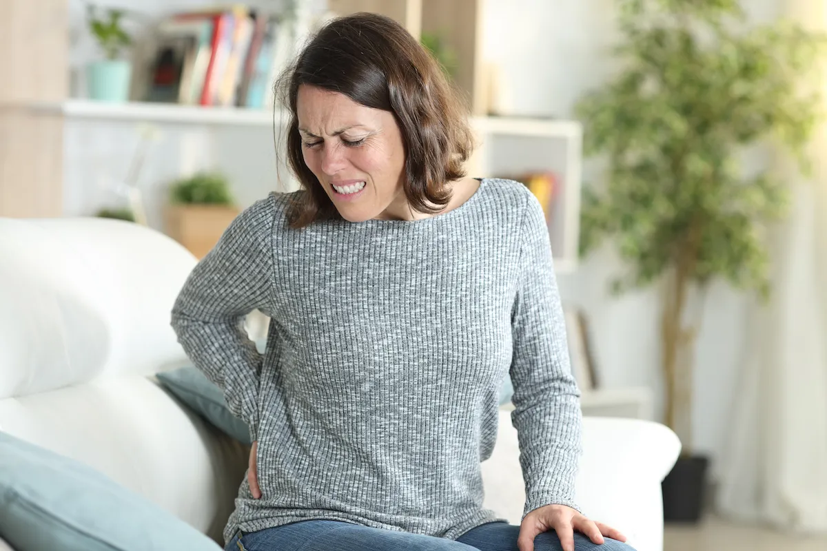 Woman on a couch wincing and holding her lower back.